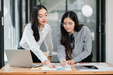 Two businesswomen working together in a modern office, analyzing documents and charts on a wooden table.