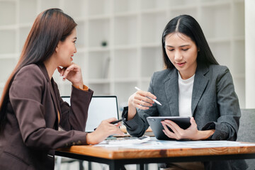 Two businesswomen in a modern office setting discussing strategy with a digital tablet and documents on the table.