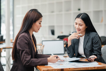 Two businesswomen discussing project details in a modern office, surrounded by laptops and documents.