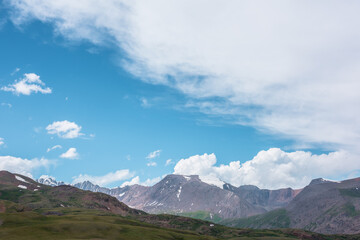 Dramatic alpine view from green hills to large rocky mountain range with glacier under clouds in blue sky. Scenic landscape with big snow mountain top. Changeable cloudy weather in high mountains.