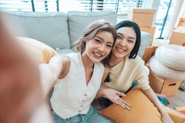 Two young Asian women smiling and happy while taking a selfie in their living room at home, looking at the camera