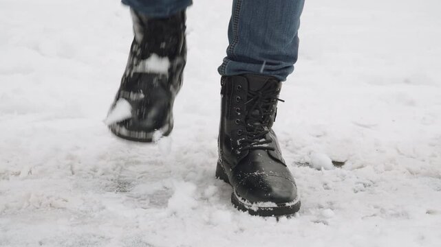 Close up of men boots on man feet in snow. Person stamps feet, shaking snow off surface of black boots during snowfall in winter. City sidewalks and nuances of walking on snowdrift.