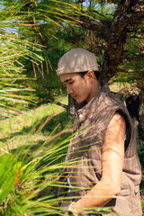 An asian solitary man wearing a stylish hat, beige clothes is standing quietly in a dense forest, surrounded by greenery and the natural beauty of the woodland environment