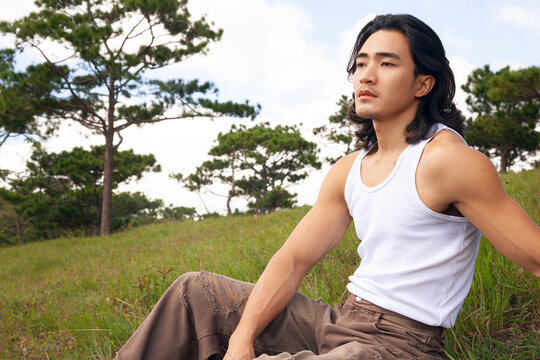 An asian handsome man with long hair wearing white tank top seat in a grassy field, surrounded by nature, soaking in the tranquility of the outdoors.