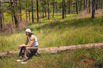 An asian handsome man with long hair wearing white tank top, beige stylist hat sits on a log in a serene field, surrounded by greenery and open space, enjoying the peaceful outdoors.