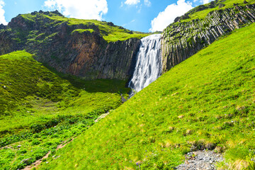 Beautiful Terskol waterfall falls from a basalt wall and is surrounded by green slopes. Caucasus,...