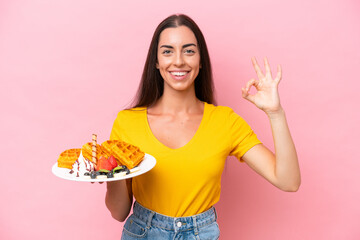 Young caucasian woman holding waffles isolated on pink background showing ok sign with fingers