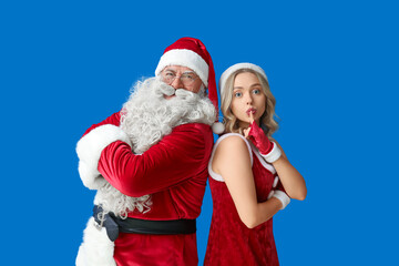 Santa Claus with crossed arms and beautiful young woman showing silence gesture on blue background
