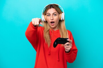 Young Uruguayan woman playing with a video game controller isolated on blue surprised and pointing front © luismolinero