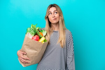 Young Uruguayan woman holding a grocery shopping bag isolated on blue background and looking up