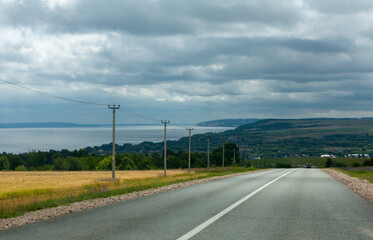 A tranquil road winds alongside the Vilga river, bordered by lush fields and distant hills under cloudy skies.