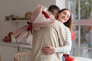 Beautiful young couple with bouquet of roses hugging and celebrating Valentine's Day in kitchen at...