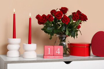 Bouquet of roses, candles, gift boxes and cube calendar with date 14 FEBRUARY on commode near beige wall at home decorated for Valentine's Day celebration