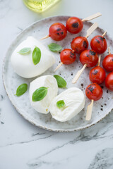 Buffalo mozzarella with grilled cherry tomatoes on wooden skewers and fresh basil, vertical shot on a white marble background, middle closeup