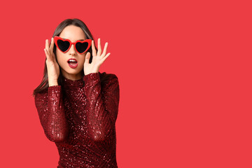 Beautiful young woman in stylish heart shaped sunglasses on red background. Valentine's Day celebration
