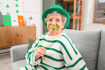 Senior woman in leprechaun hat with paper mustache sitting on sofa at home. St. Patrick's Day celebration