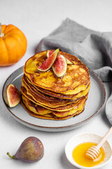 Stack of pumpkin pancakes with honey and figs in a plate on light background with fresh fruits and vegetables.
