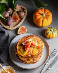 Stack of pumpkin and pancakes with honey and figs on a dark background with fresh fruits and vegetables.