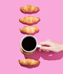 Hand holding a cup of espresso coffee on a pink background with shadow and fresh mini croissants.
