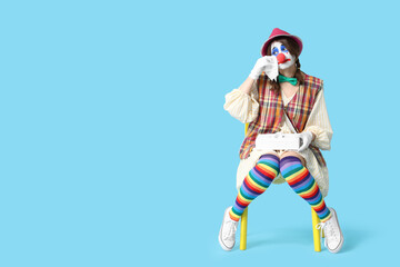 Young woman dressed as sad clown crying on chair against blue background. April Fool's day celebration