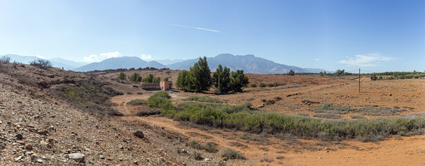 Moroccan countryside scene: red earth, sparse vegetation, a pink house, and distant mountains under a serene sky.