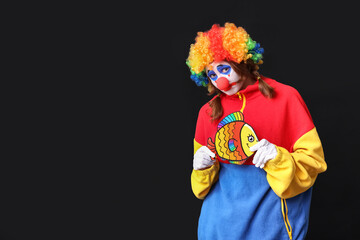 Young woman dressed as sad clown with paper fish on black background. April Fool's day celebration