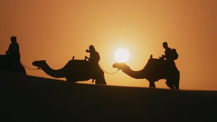 Silhouette of tourists riding camels on top of a dune in a safari desert tour during sunset - Powered by Adobe