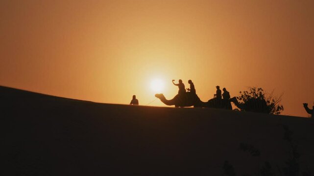 Silhouette of tourists riding camels on top of a dune in a safari desert tour during sunset