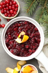 Tasty cranberry sauce in gravy boat, berries, orange peels and fir branches on grey table, flat lay