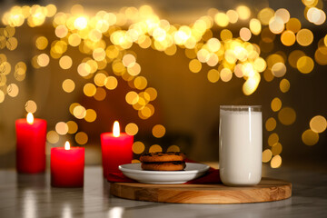 Glass of milk, tasty chocolate chip cookies and burning candles on table against blurred Christmas lights. Bokeh effect