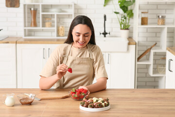 Beautiful young woman dipping fresh strawberry into bowl with melted chocolate in kitchen