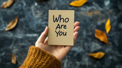 Close-up of a hand holding a card that says 'Who Are You' amid scattered autumn leaves.