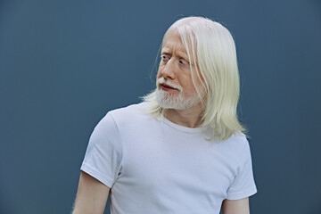 Elderly man with long white hair and beard looking to the side against a simple blue background, wearing a plain white t shirt, capturing a moment of contemplation