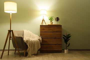Interior of living room with glowing lamps, armchair and commode in evening