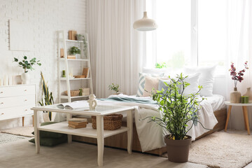 Interior of light bedroom with tables, shelf unit and plants