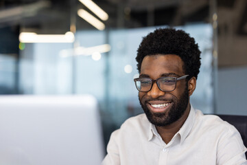 African American businessman smiling while working on computer in modern office. Glowing screen reflects focus, productivity. Professional attire highlights engagement, technology use.