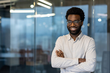 Obraz premium African American businessman in modern office with crossed arms, wearing glasses and shirt. Confident expression reflects professionalism and success in corporate environment, radiating positivity.