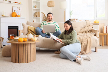 Happy young man using laptop while his wife reading magazine at home on autumn day