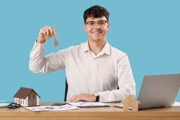 Male real estate agent with key from house at table on blue background