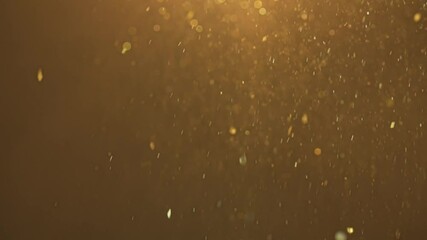 Many sparkling glitters showering floating in light on empty golden background with light from above.