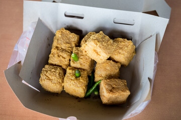 Close-up of crispy, golden-brown fried tofu in a cardboard container with green chili peppers. The texture and savory appeal are highlighted.