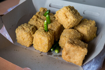 Close-up of crispy, golden-brown fried tofu in a cardboard container with green chili peppers. The texture and savory appeal are highlighted.