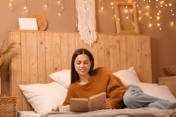 Beautiful young happy woman with cup of cacao reading book on bed at home in evening
