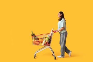 Beautiful young happy woman with full shopping cart on yellow background