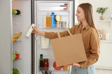Young woman putting yoghurt from shopping bag into fridge in kitchen