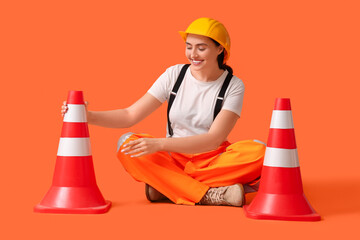 Young female construction worker with traffic cones sitting on orange background. Woman in male...