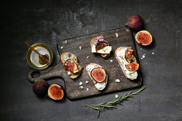 Wooden board of delicious fig bruschetta with different cheese and bowl of honey on black background