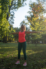 Fit, athletic woman stands in a park, enjoying the late summer breeze. With her arms wide open and a bright smile on her face, she radiates positivity and energy while exercising outdoors.