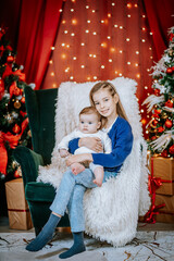 Obraz premium A young girl holds a baby on a cozy chair surrounded by festive Christmas lights, decorated trees, and wrapped gifts in a warm holiday setting.