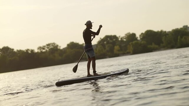 Surfer Balancing On SUP Board In River Against Urban Skyline. Summer Aquatic Activity